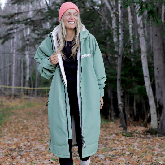 Woman wearing a long green coat with a brand logo, standing in a forest.